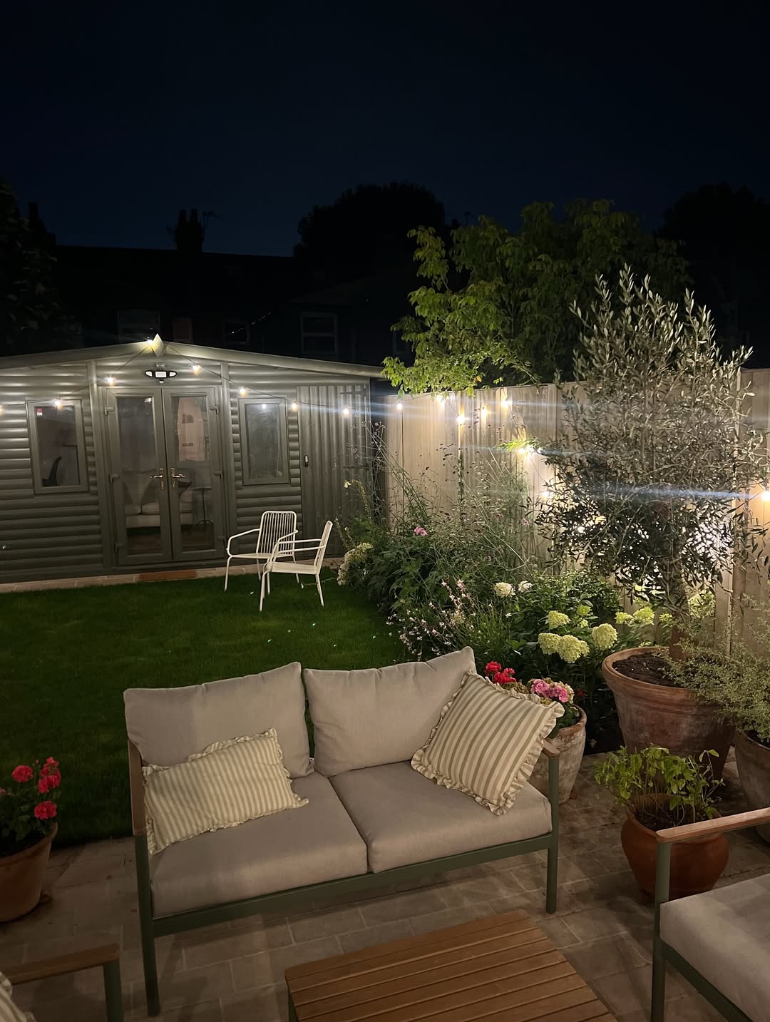 Backyard patio at night with string lights, a lit garden room and an olive tree in a terracotta pot