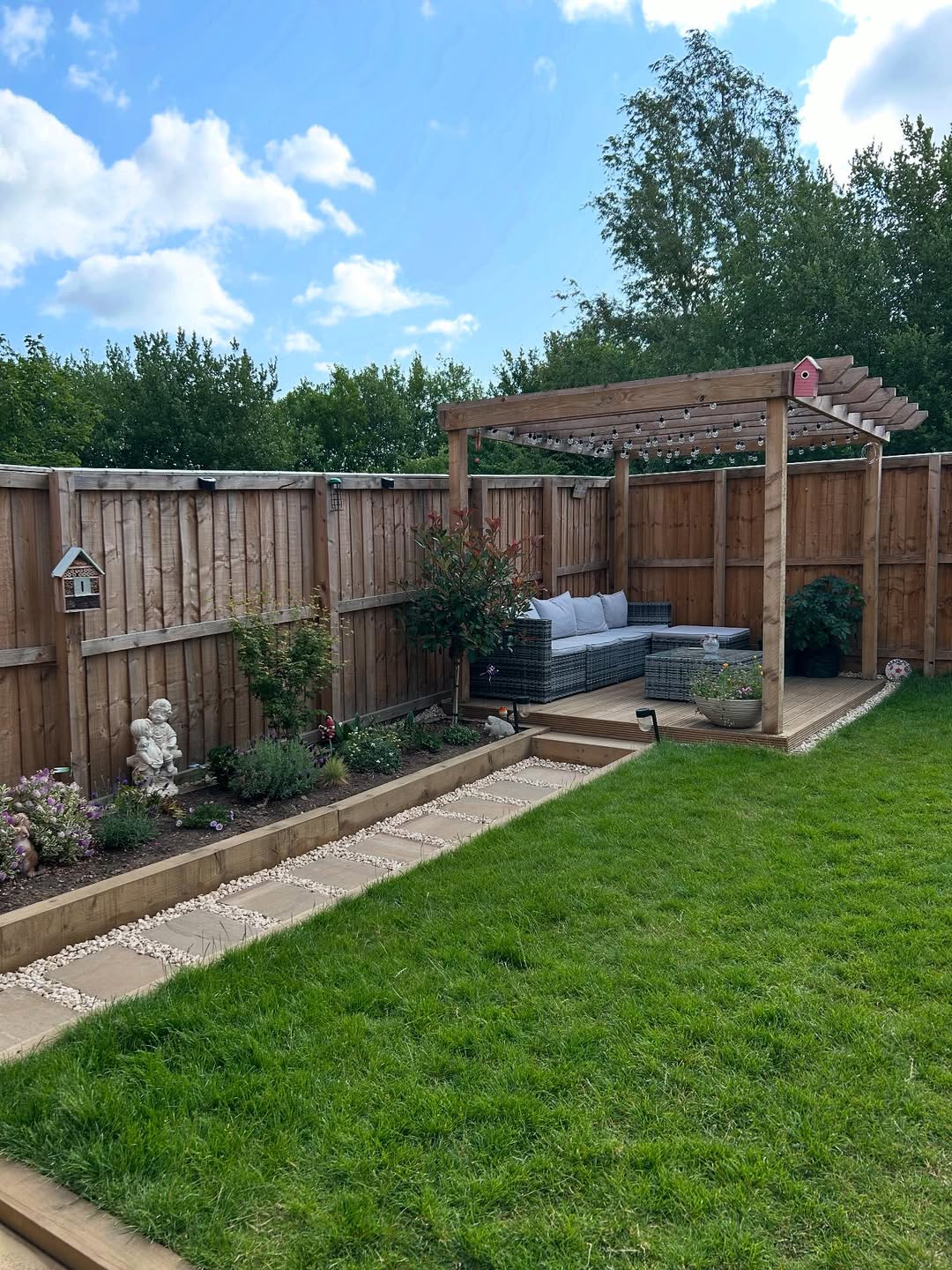 Wooden pergola over a decked seating area with rattan sofa at the end of a narrow lawn