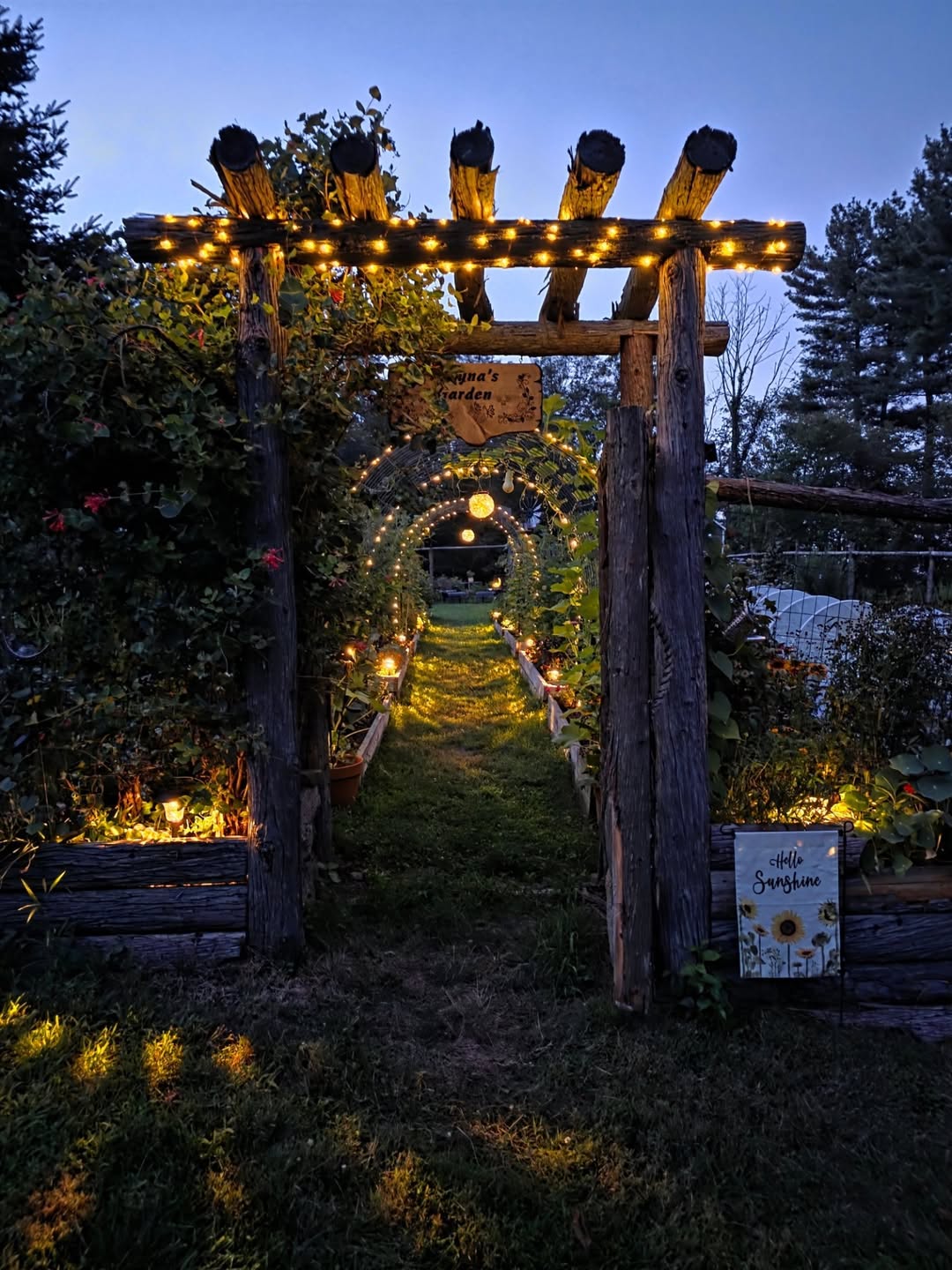 Rustic log arch strung with fairy lights over a grass path into a vegetable garden at dusk